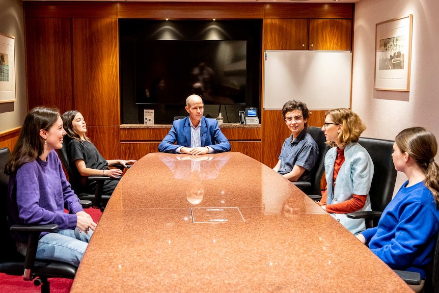 A older man in a boardroom with five teenagers