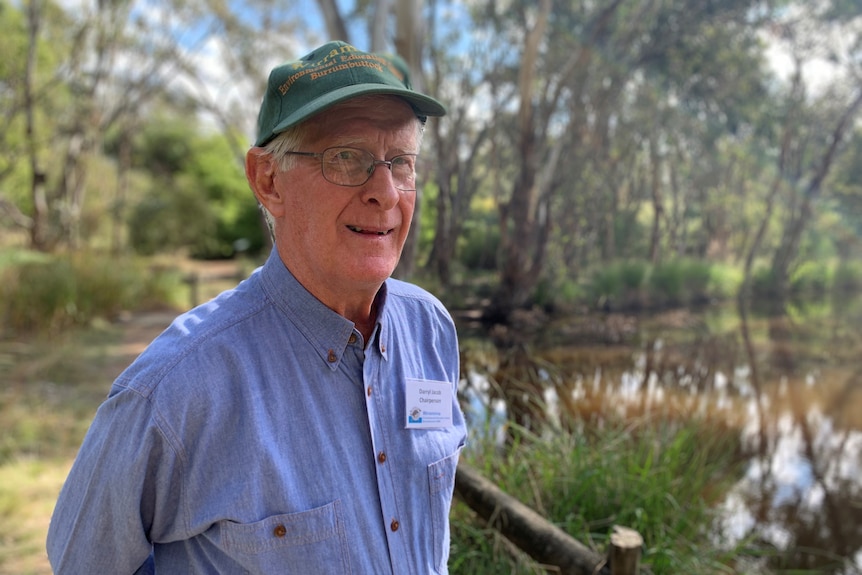 An older man with greying hair, green cap, glasses, blue shirt, stands in a bush reserve, with dam behind him.  Looks serious.