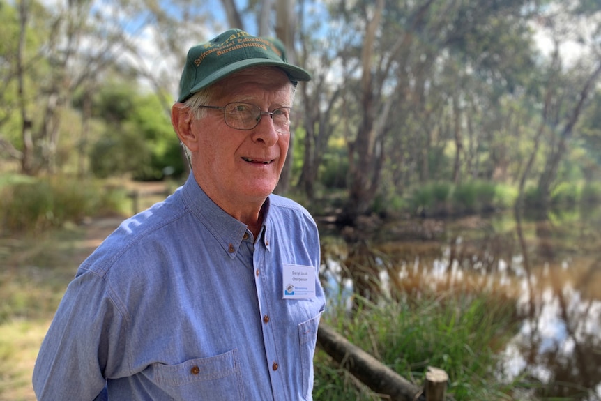 An older man with greying hair, green cap, glasses, blue shirt, stands in a bush reserve,  with dam behind him. Looks serious.