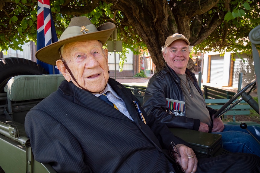 two men, one elderly wearing an army slouch hat and another man behind, sit on the front seat of a World War II jeep.