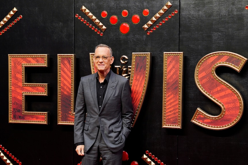 Tom Hanks in a dark grey suit stands in front of a red ELVIS banner