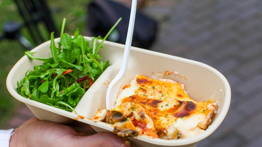 A vegetarian lasagne in a takeaway container with a salad beside it and a fork stuck into it