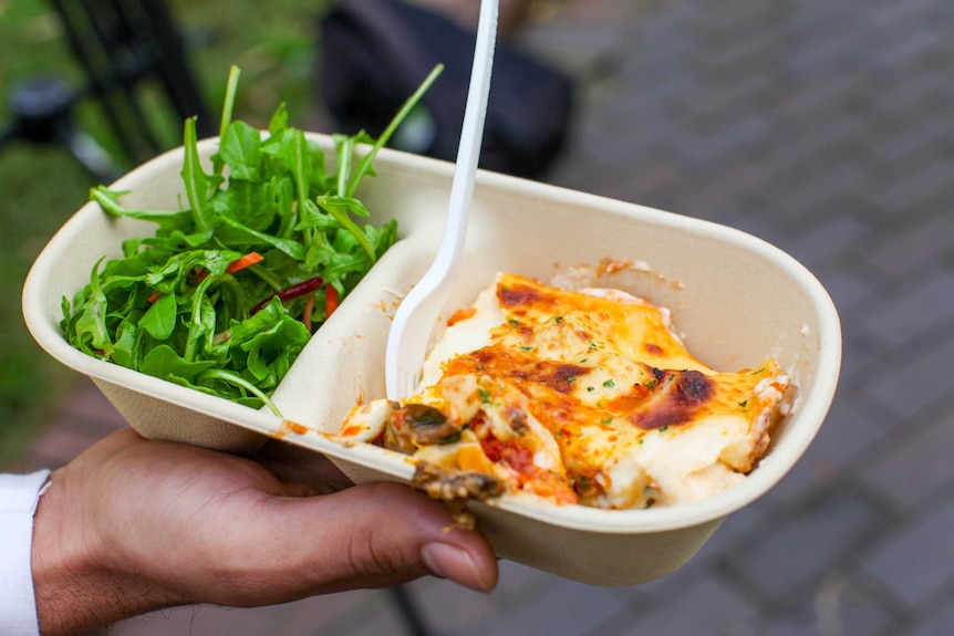 A vegetarian lasagne in a takeaway container with a salad beside it and a fork stuck into it