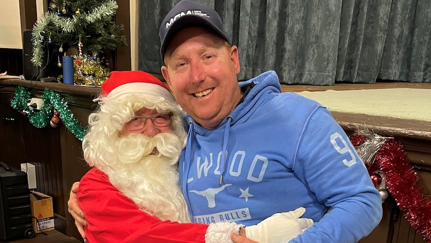 A man in a Santa outfit hugs a man in his 30s on his lap.