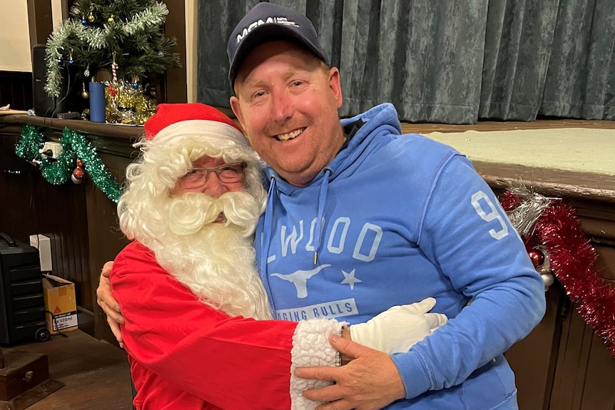 A man in a Santa outfit hugs a man in his 30s on his lap.