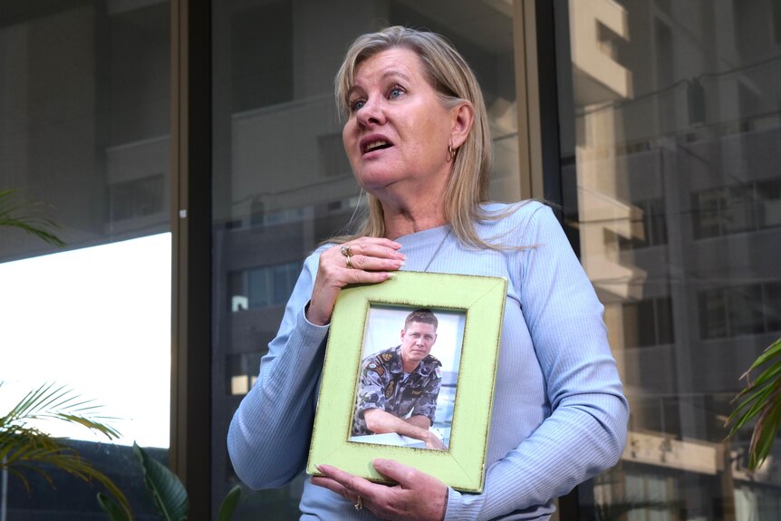 A woman looking anguished holding a picture of a man in military uniform in a frame.