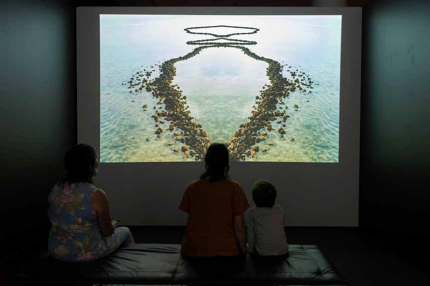Three people sitting on a settee watching a video on a large screen, inside a dark room inside an art gallery.
