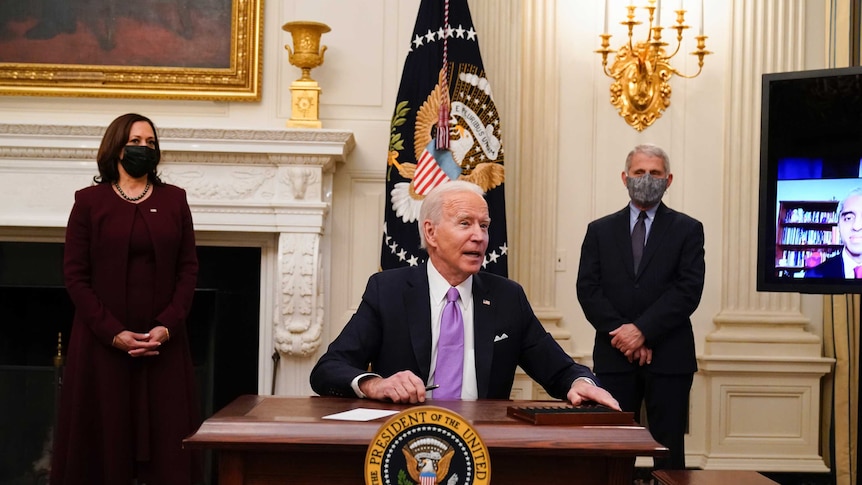 A man wearing a suit and purple tie sits at a desk with the presidential seal on it flanked on either side by a woman and man.