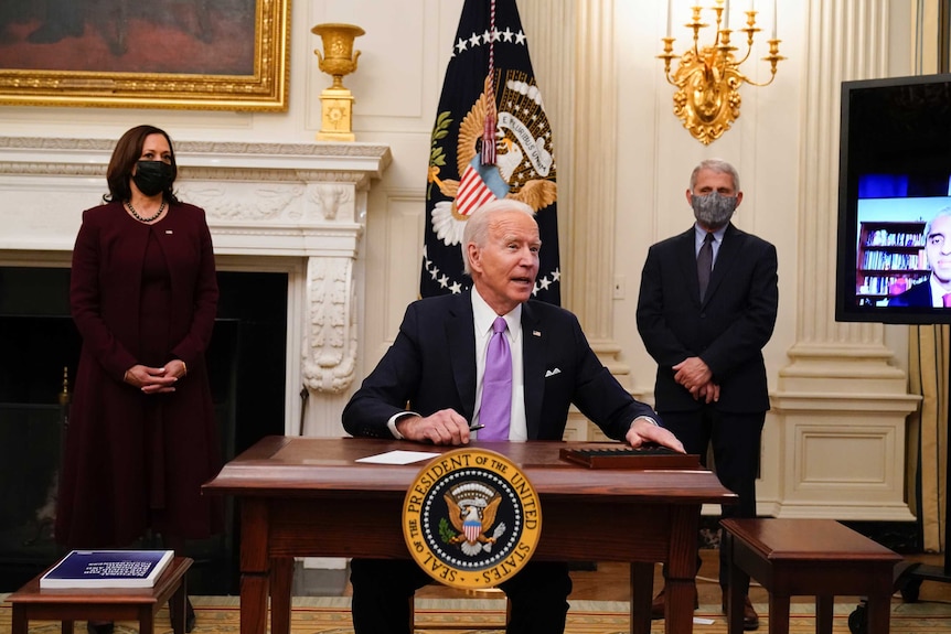 A man wearing a suit and purple tie sits at a desk with the presidential seal on it flanked on either side by a woman and man.