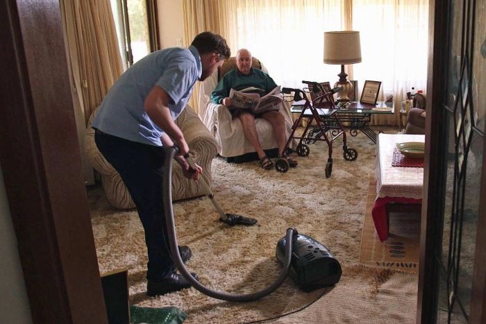 A man vacuums a carpet in a loungeroom as an elderly man sits in an armchair