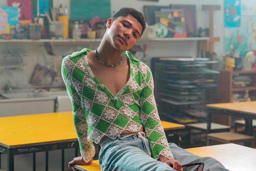 James Majoos wears a green and white crochet top and sits on a desk in a classroom, while looking to the camera.
