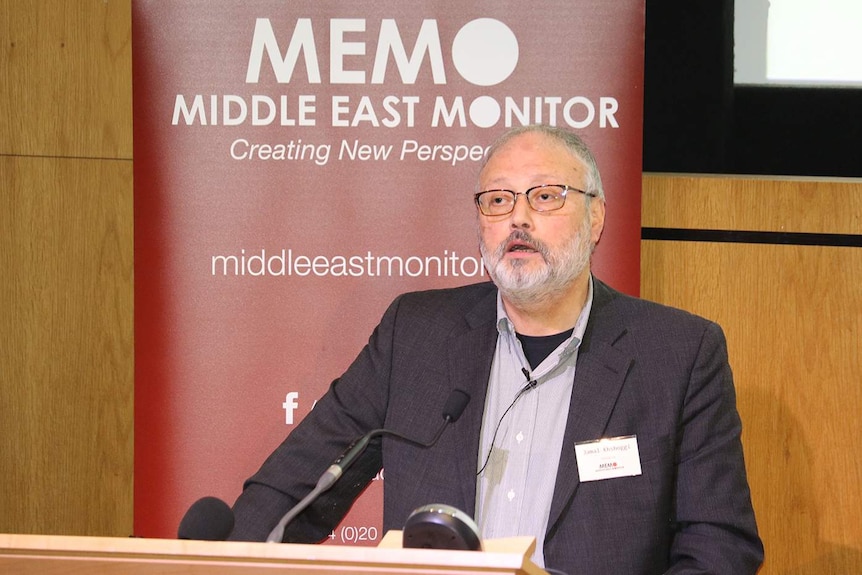 A bearded man with grey hair and a beard dressed in a shirt and suit jacket speaks from a podium