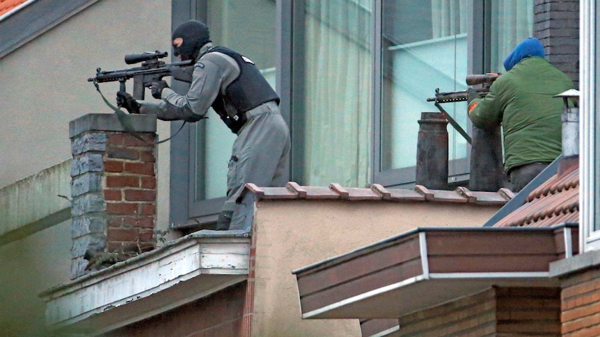 Photo from street level of two masked police standing behind chimneys on a rooftop, pointing their guns.