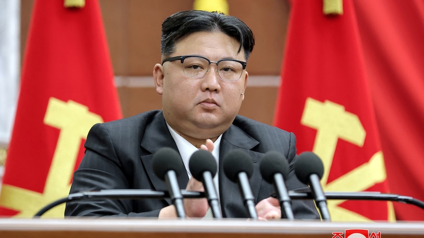 A heavy-set Korean man with glasses and a peculiar headcut sits behind a row of microphphones in front of two red and gold flags