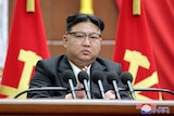 A heavy-set Korean man with glasses and a peculiar headcut sits behind a row of microphphones in front of two red and gold flags