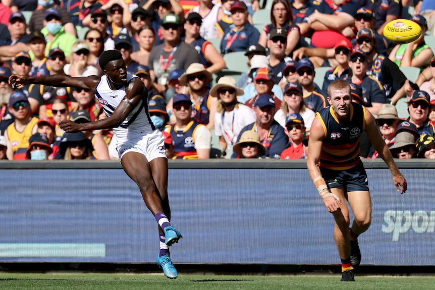 An AFL footballer looks downfield as he snaps a kick across his body toward goal as a defender watches. 