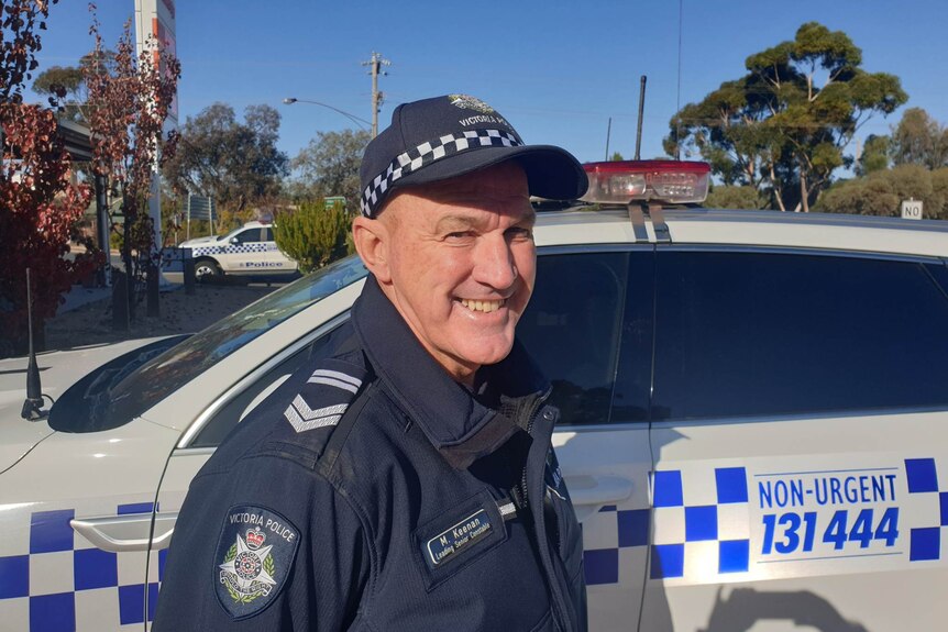 Man in police uniform stands next to a white and blue police car, smiling.