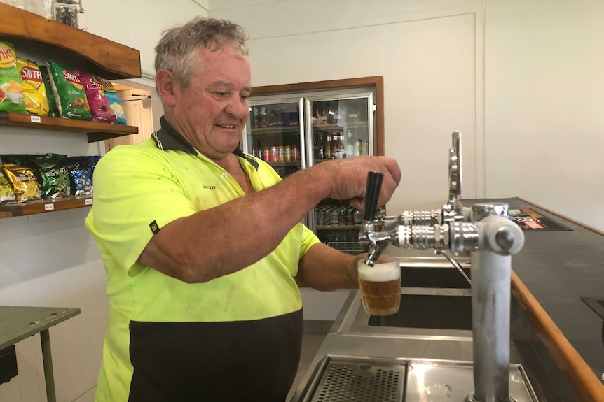 A man pouring a beer in a pub. chips behind him. drink fridge to the right of him