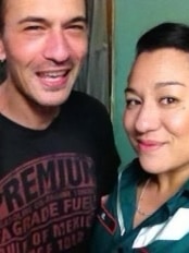 A man in a black t-shirt grins as he stands next to a dark-haired woman wearing a green SA Ambulance uniform