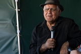 Elderly Indigenous Australian man wearing a black bowler hat and black shirt holds a microphone inside a green tent.
