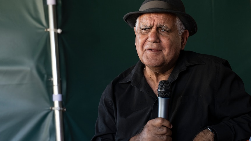 Elderly Indigenous Australian man wearing a black bowler hat and black shirt holds a microphone inside a green tent.
