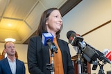 A woman wearing a black suit and orange shirt speaks to microphones while standing in front of two men