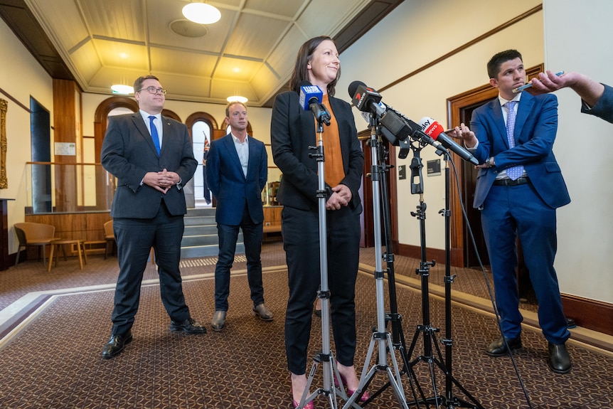 A woman wearing a black suit and orange shirt speaks to microphones while standing in front of two men