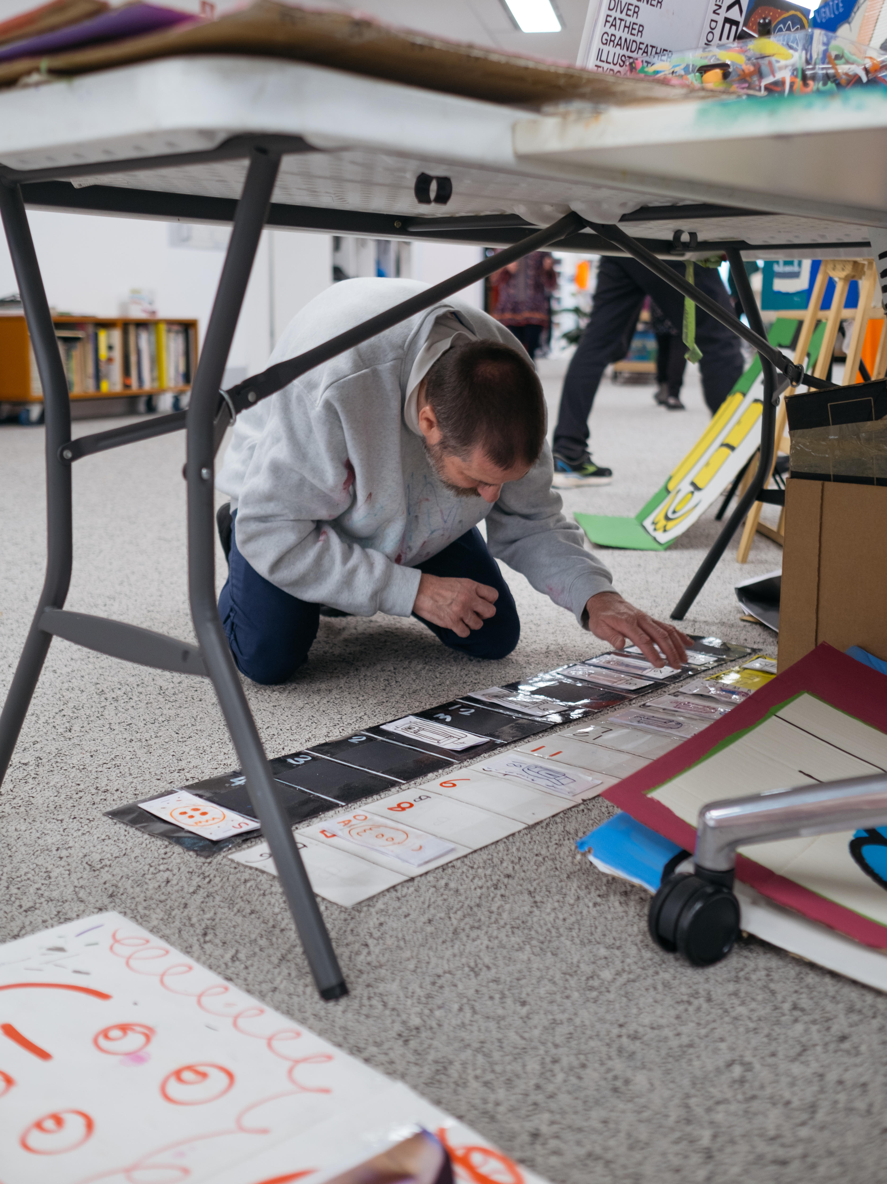 Artist Thom crouches under his working table in the studio, working on an artwork on the floor