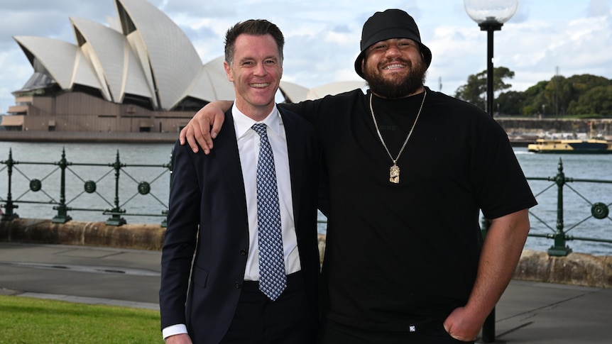 two men smiling standing outside next to the harbour in  front of the opera house one man wears a hat