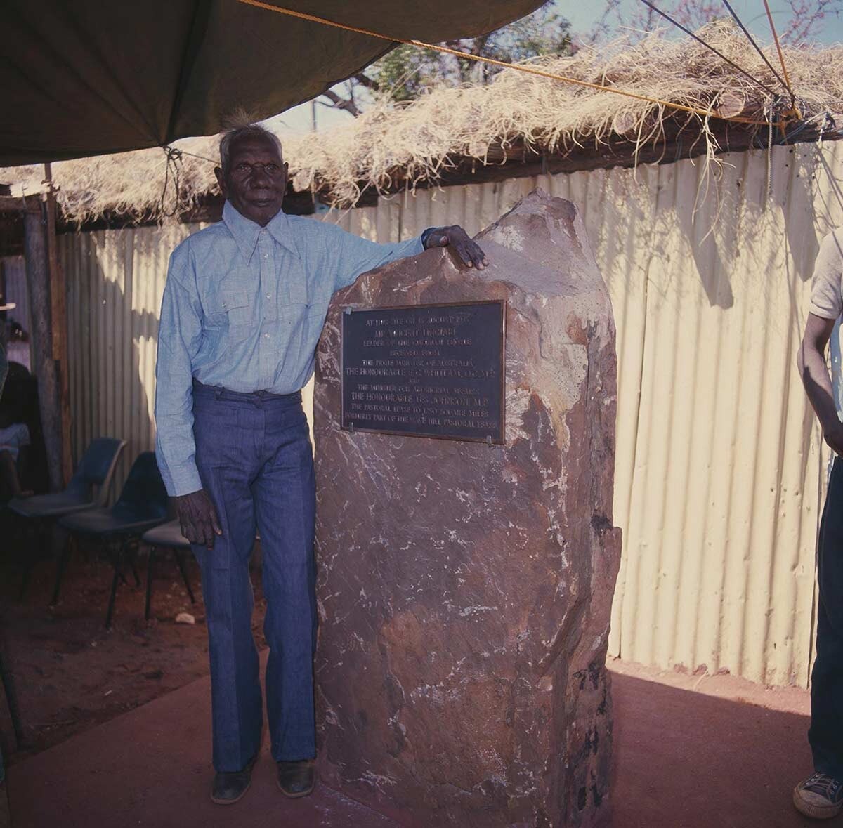 An Aboriginal man in a blue button up shirt leaning against a big rock with a plaque on it. Photo is dated.
