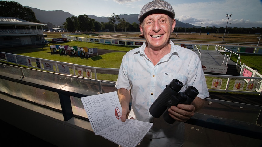 A man wearing a flat cap, holding a form guide and binoculars, in the stands at a racetrack.