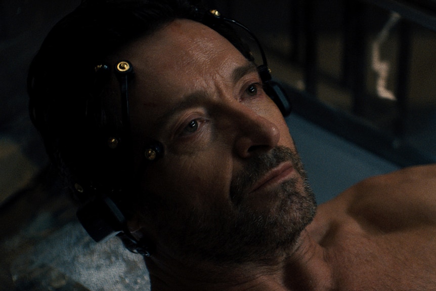 A close up on a dark and brooding Hugh Jackman. He is lying down in a high-tech pod-like machine and is intently focused.