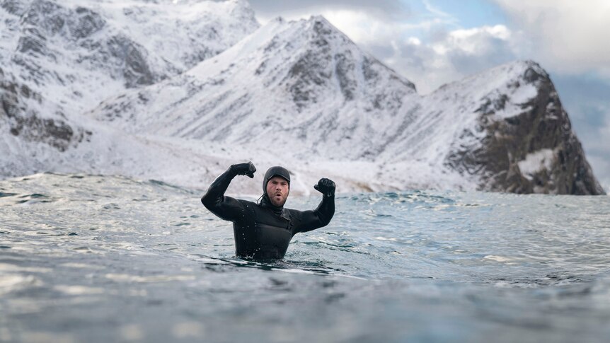 Chris Hemsworth wearing a black wetsuit while swimming in water with snowy mountains in the background