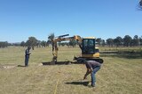 Two forensic police officers examine a grassy paddock in Armidale with an excavator used to dig the ground.