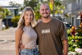 A Caucasian woman and man stand arm in arm smiling outside of a house on a sunny day.