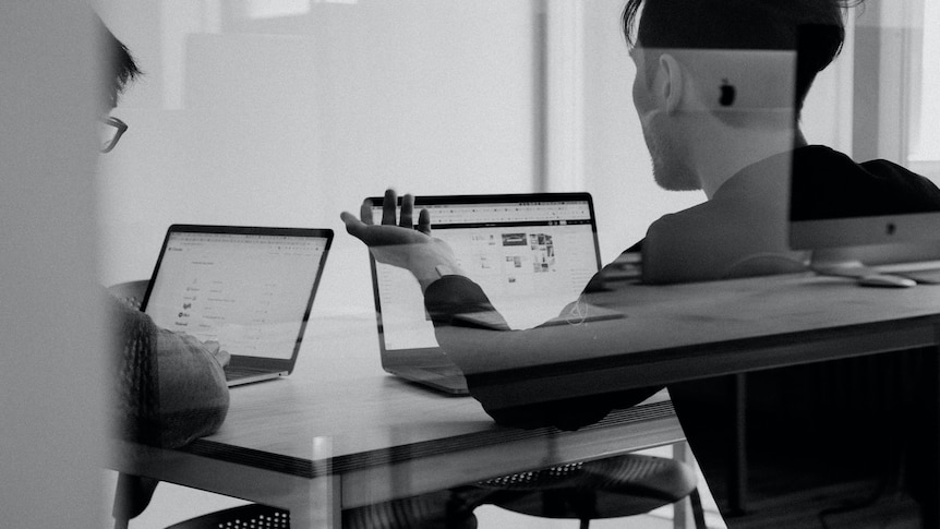 a monochrome of the backs of two people in an office looking concerned about what's on their screen.