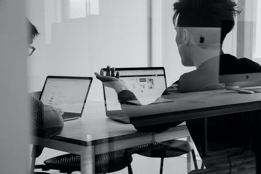 a monochrome of the backs of two people in an office looking concerned about what's on their screen.