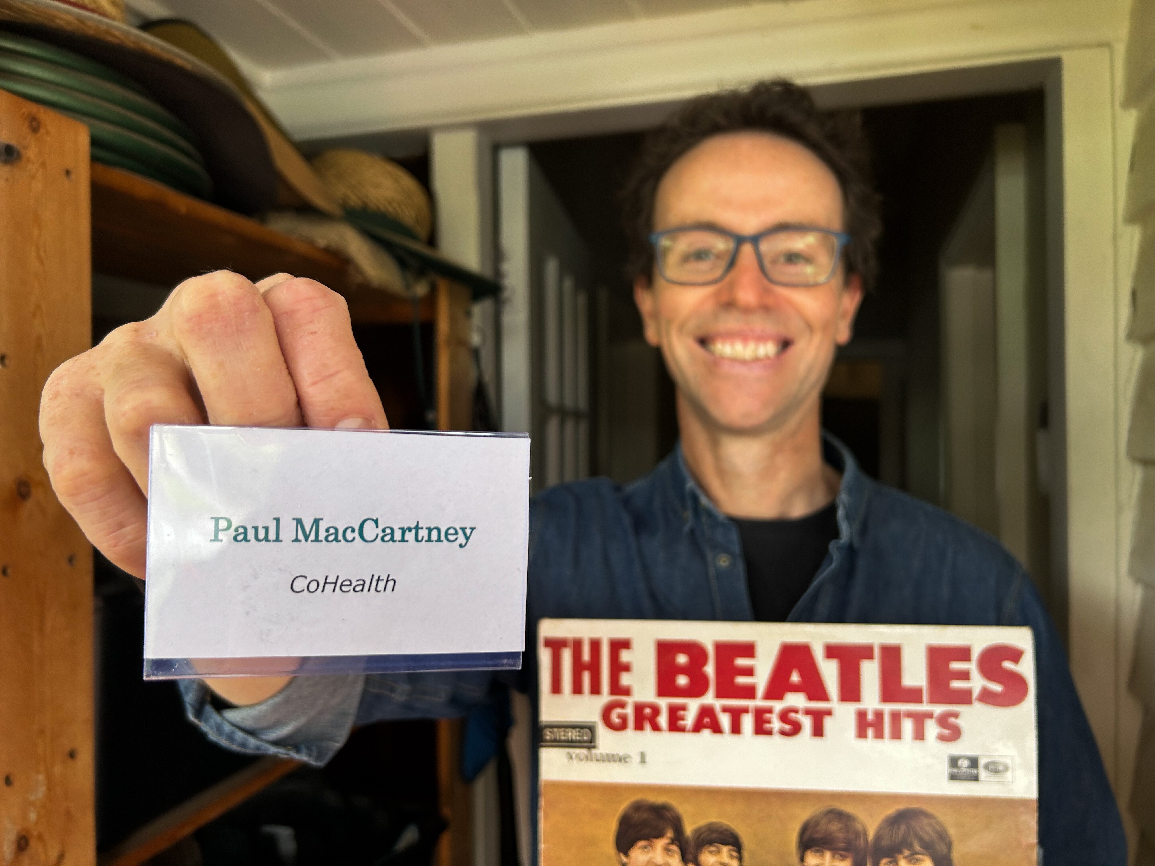 Man with glasses stands at front door smiling, holding a name badge reading Paul MacCartney CoHealth and a Beatles record