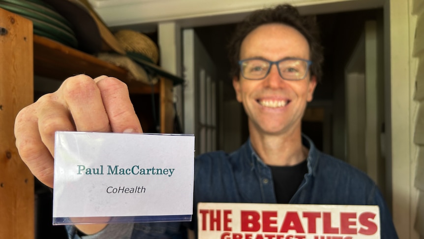 Man with glasses stands at front door smiling, holding a name badge reading Paul MacCartney CoHealth and a Beatles record
