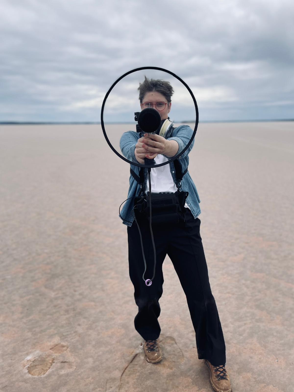 Woman standing on a salt plain pointing microphone with clear disc around it at the camera.
