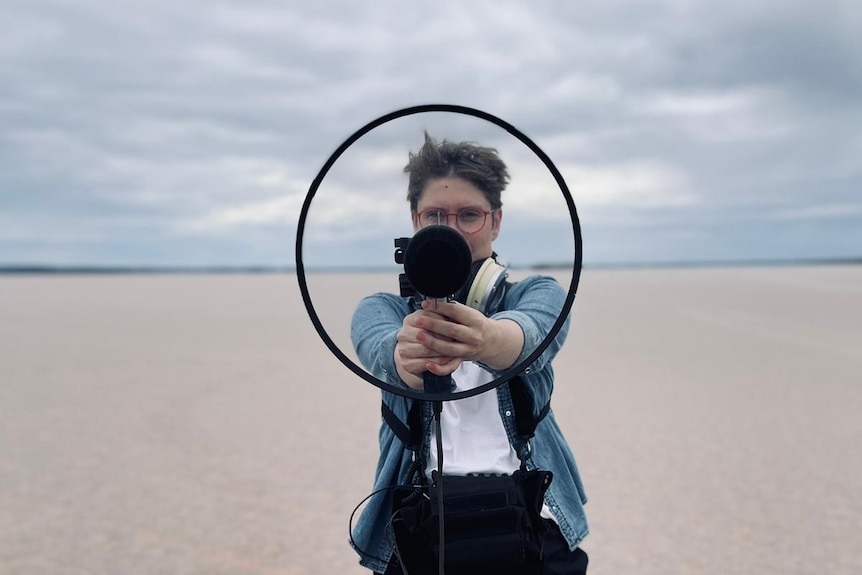 Woman standing on a salt plain pointing microphone with clear disc around it at the camera.