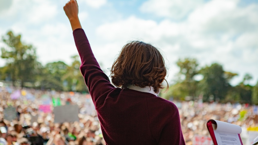 A view behind of a student on stage with short brown hair raising her fist in the air, a large crowd is blurred in front of her.