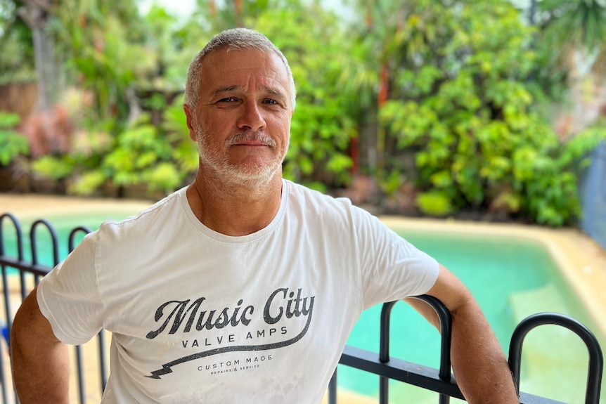 A greying man looks at the camera while leaning against a fence with a pool behind him