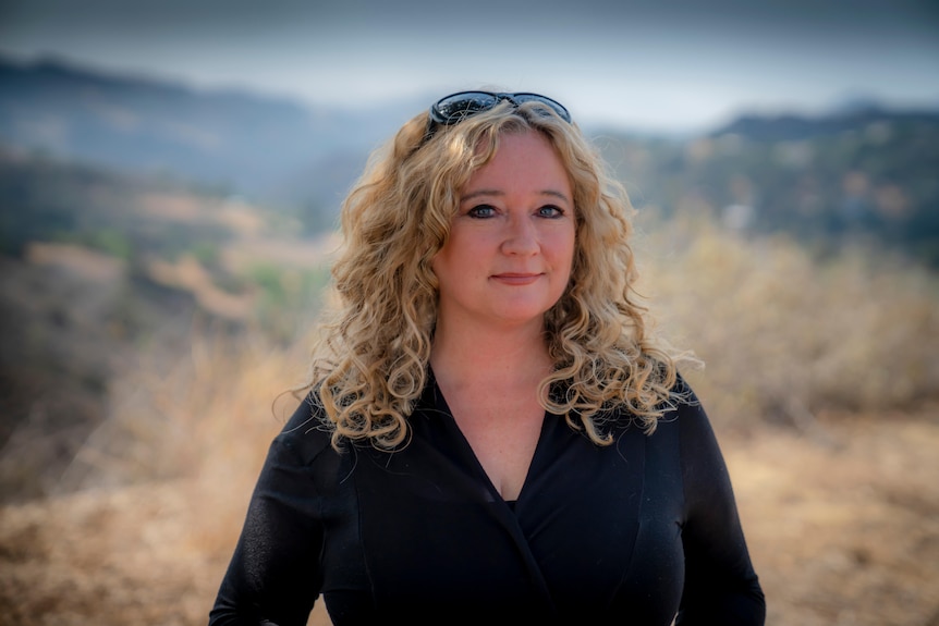 A middle-aged woman with blonde curly hair stands outside.