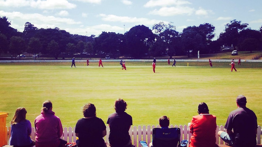 Spectators watch a cricket match on an oval.