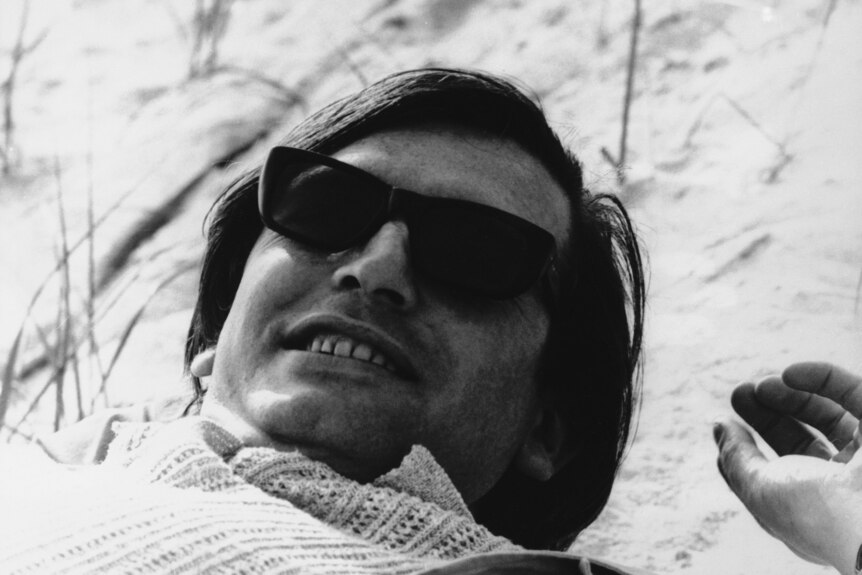 A black and white photo shows a man in his 30s wearing sunglasses lying on his back in sand.