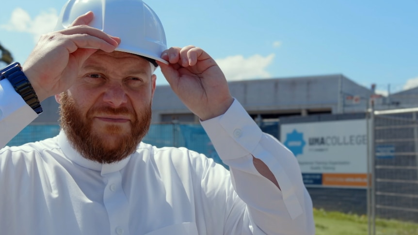Man with fair skin and orange beard wears white shirt and white hard hat in front of contstruction site