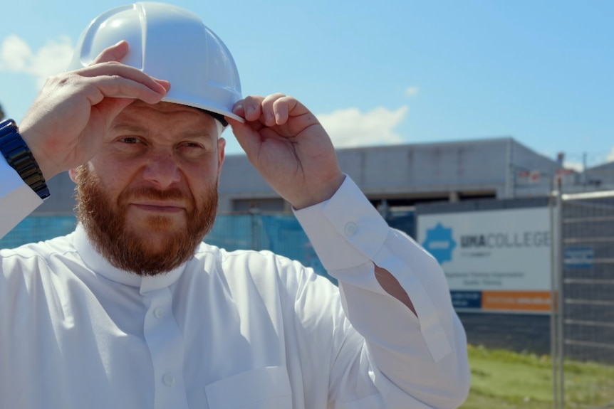 Man with fair skin and orange beard wears white shirt and white hard hat in front of contstruction site