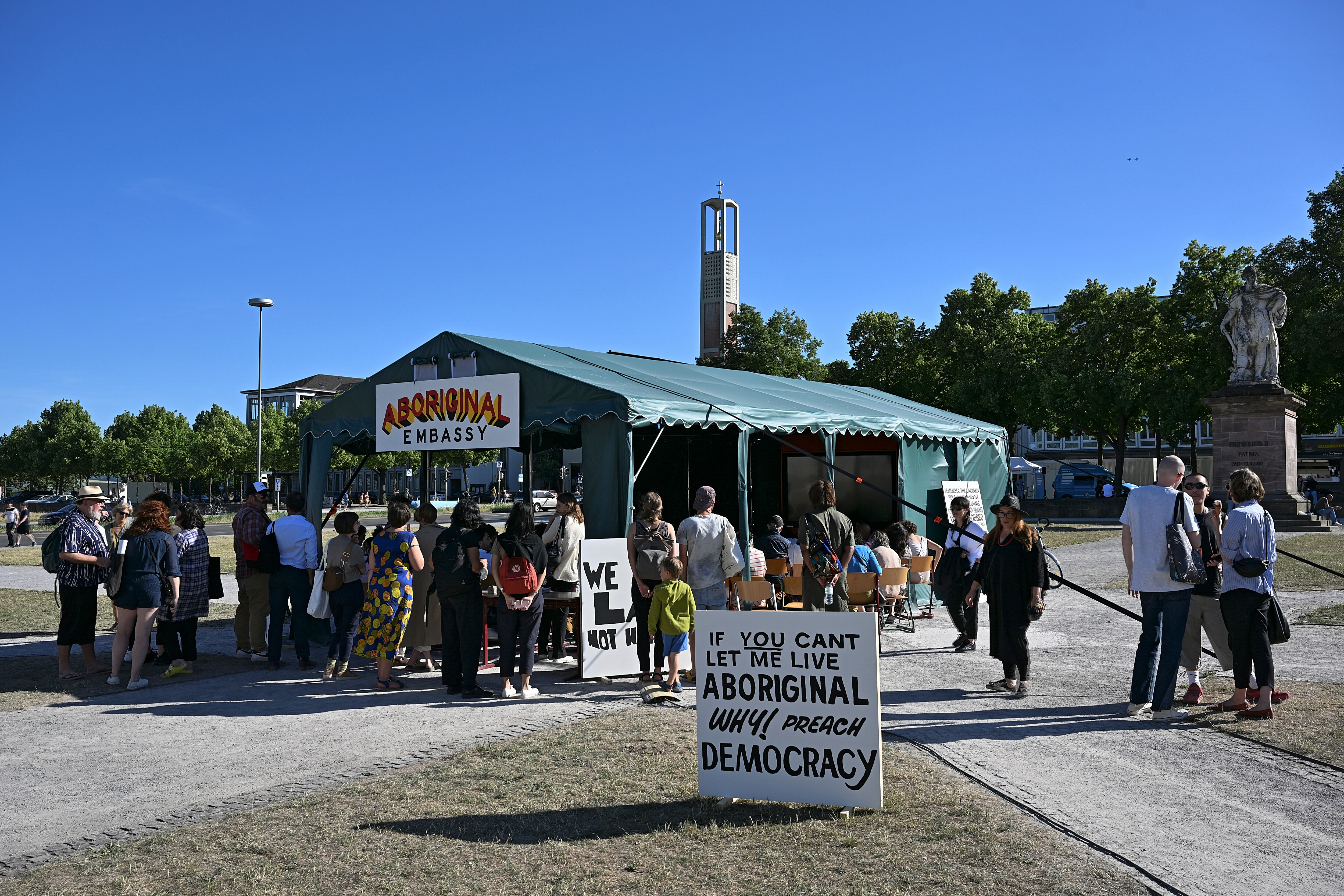 A group of people queue outside a large green tent bearing a yellow, red and black sign that says "Aboriginal embassy" in a park