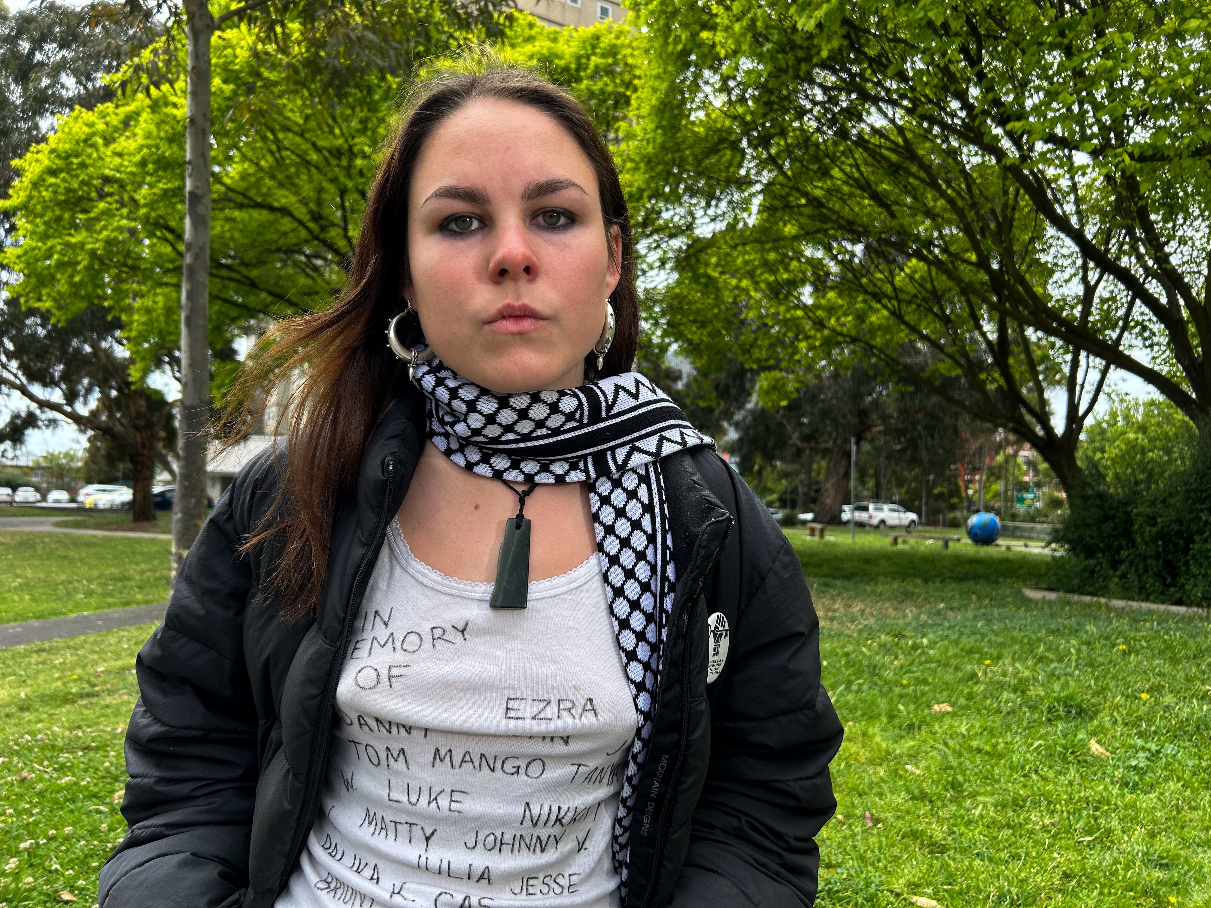 Aaneke has long brown hair, hoop earrings, a black and white scarf, black puffer, necklace and white singlet with names on it. 